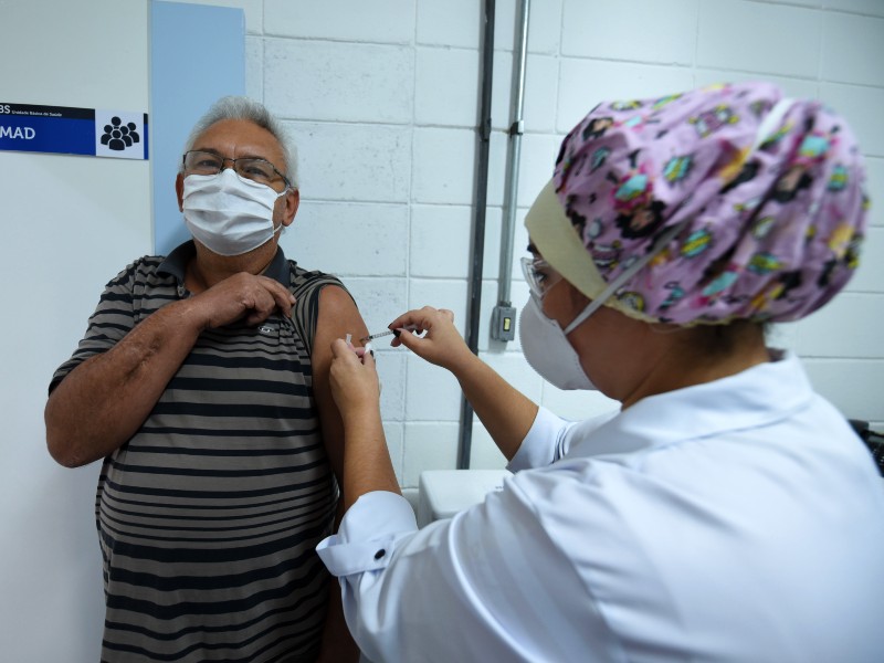 Foto de vacinação contra a Covid-19 para maiores de 60 anos. Profissional da saúde aplica a vacina no braço de um homem, que está de máscara, óculos e olhando para a frente com uma expressão alegre. Eles estão numa sala de vacinação com paredes brancas, dentro de uma Unidade de Saúde